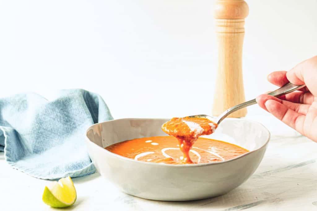 healthy vegan tomato coconut soup with curry in bowl with hand raising spoonful of soup